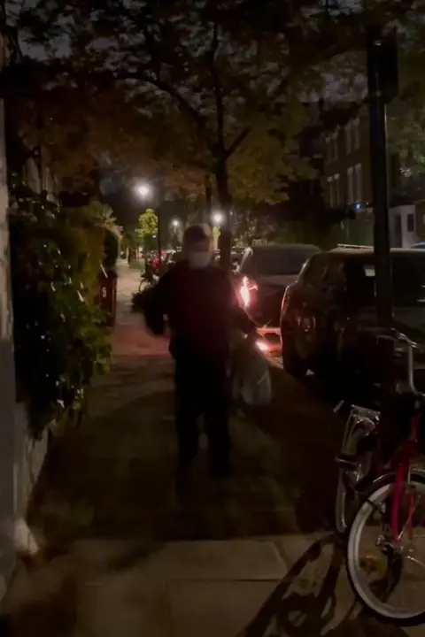 Metropolitan Police A figure dressed in dark clothing and a white face mask stands on a pavement in the dark, next to him is a bicycle and a car, behind him another car appears to be on fire.