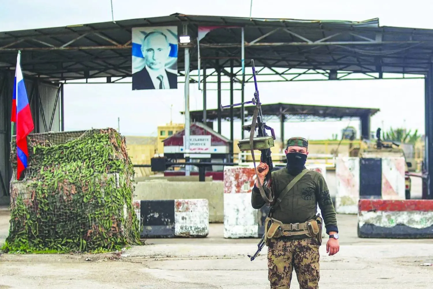 A Syrian member of the military operations looks on as he stands at an inspection checkpoint for incoming vehicles before a Russian flag and Russian soldiers behind manning the entrance of the Russian-leased Syrian military base of Hmeimim in Latakia province in western Syria on December 29, 2024. (AFP) 