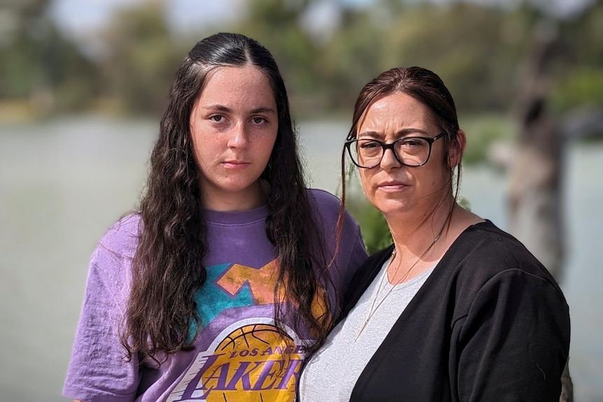 A teenager and her mother, both dark-haired, stand outside, looking solemn.